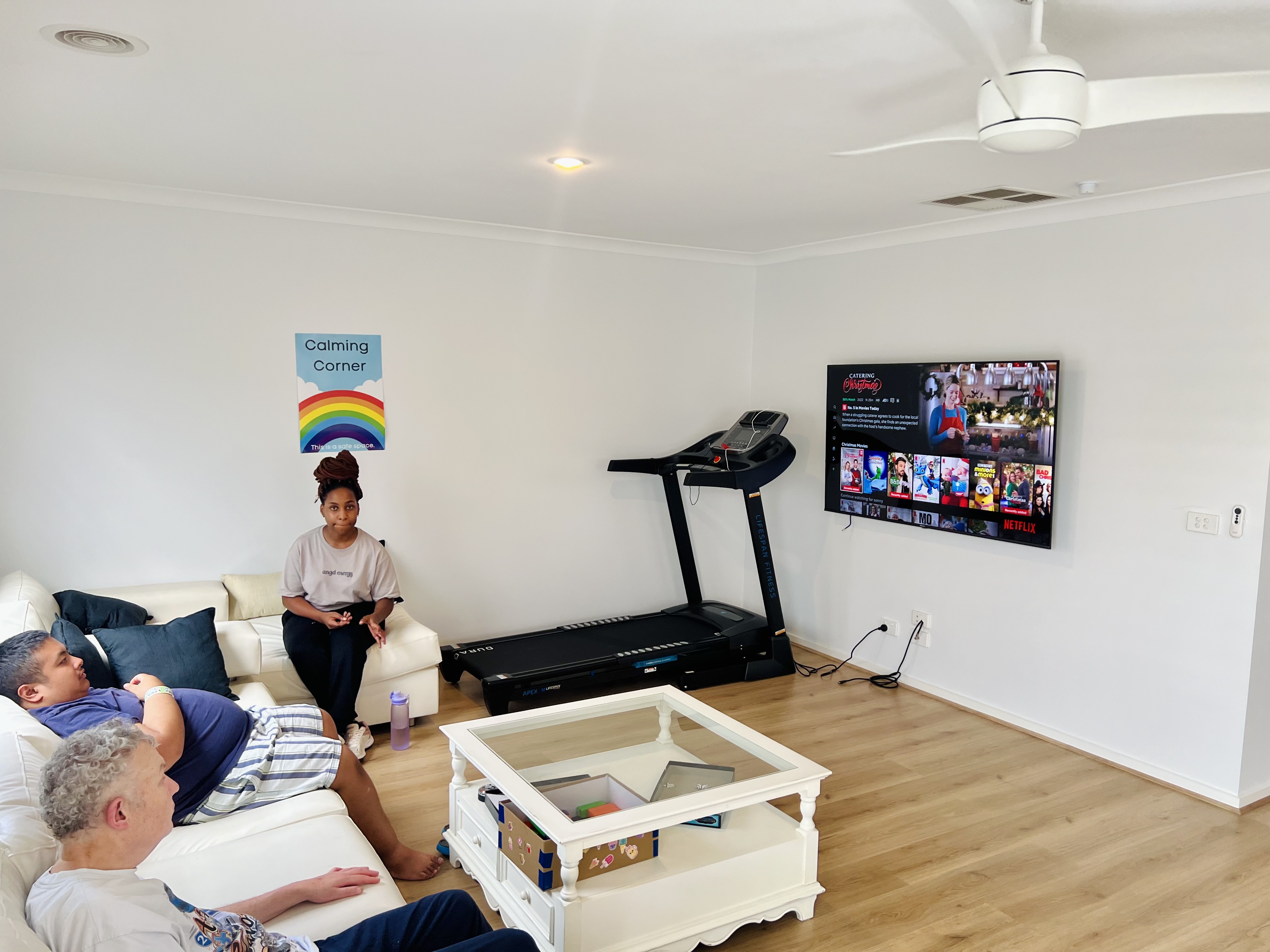 A care worker and two men are sitting on a sofa watching a wall mounted TV. There is a treadmill in the corner of the room to cater for independent living.