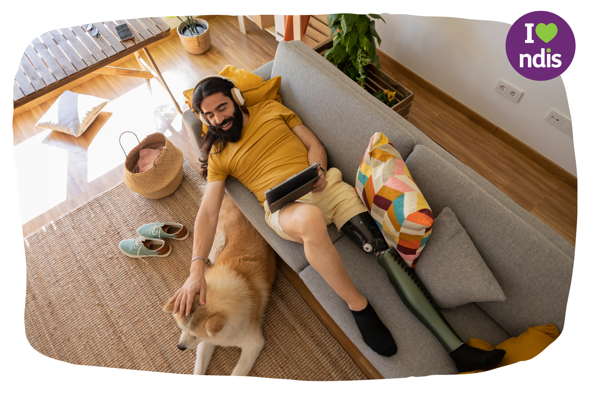 A man with a prosthetic leg is laying on his sofa with headphones on and an ipad in his hand. With his other hand he is petting his dog on the floor beside him.