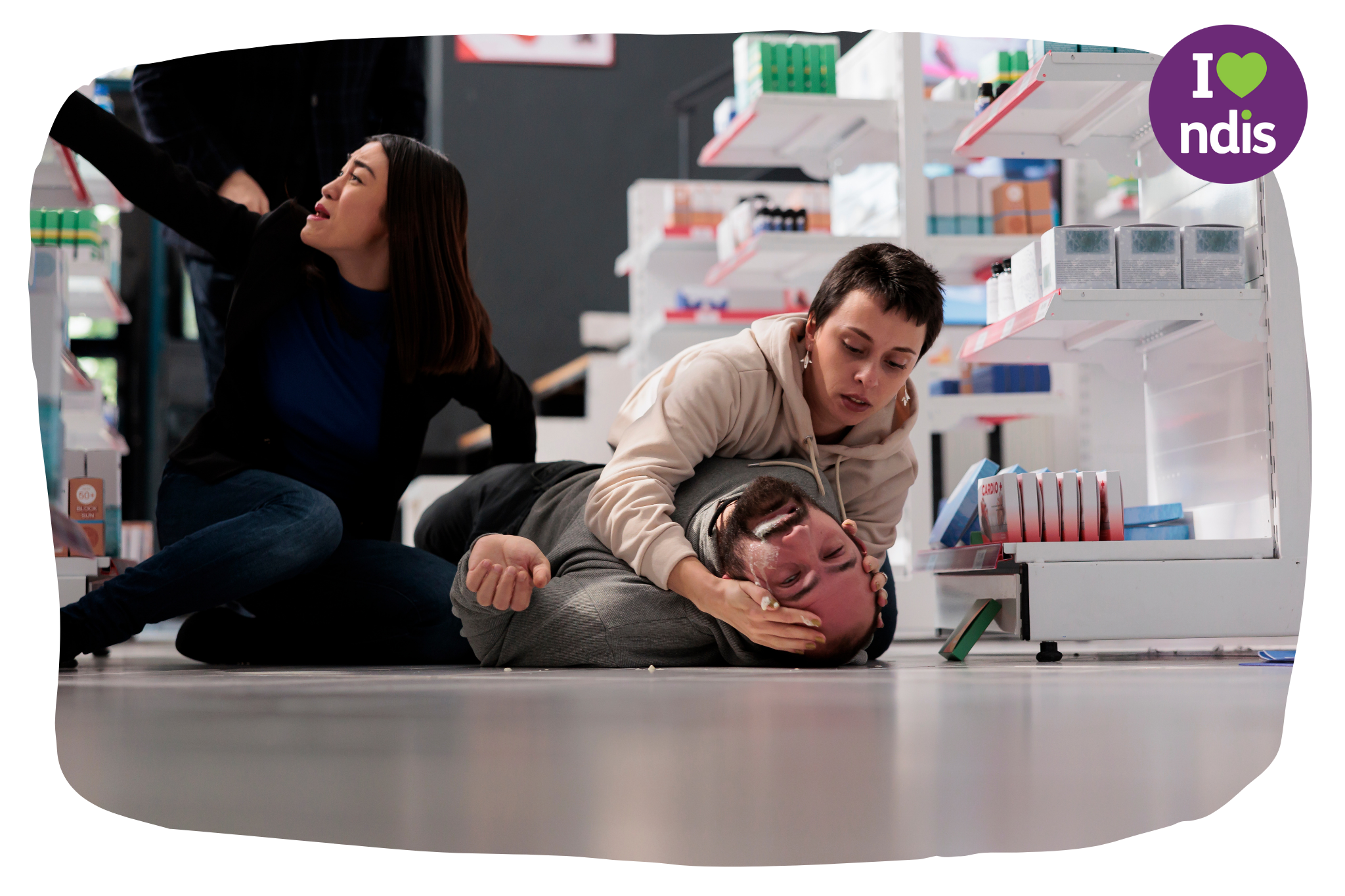 Two woman are tending to a man on the floor in a chemist. The man is having an epileptic fit.