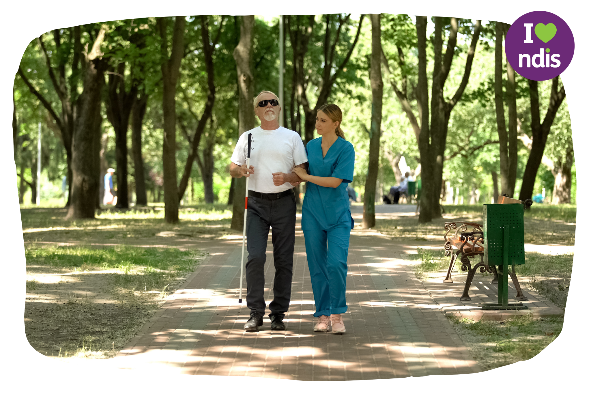 A woman in a blue nurse uniform is walking in a park holding the arm of and assisting a man who is blind.